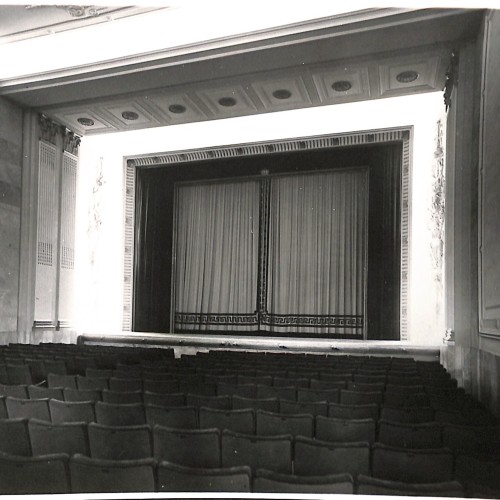 Fotografía: Vista interior del patio de butacas y embocadura del escenario/pantalla de una sala de cine o teatro de época.