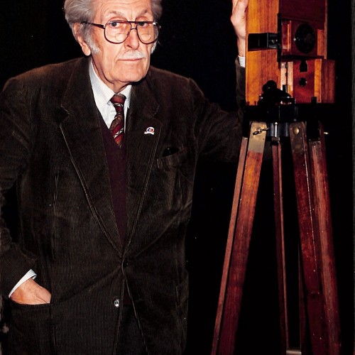 Retrato institucional de Tomás Mallol junto a una cámara cinematográfica primitiva de madera sobre trípode.