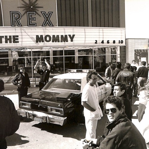 Fotografía en blanco y negro, escena exterior frente al cine REX con patrulla de policía y multitud